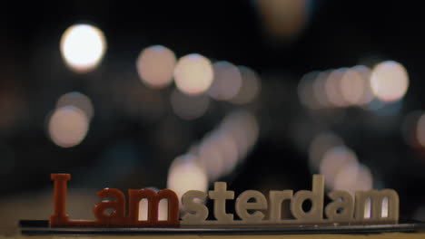 Blick-Auf-Die-Kleine-Plastikfigur-Der-Iamsterdam-Buchstabenskulptur-Auf-Der-Brücke-Vor-Verschwommenem-Stadtbild-In-Amsterdam,-Niederlande