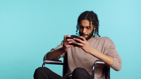 Man-with-disability-in-wheelchair-playing-videogames-on-cellphone,-studio