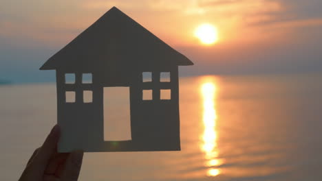 Hand-holding-cut-paper-house-against-of-sea-at-sunset