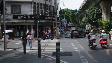Las-Personas-Que-Cruzan-Un-Carril-Peatonal-Durante-La-Señal-De-Paseo-Verde-En-Sukhumvit-26,-Mientras-Que-Las-Motocicletas-También-Aprovechan-Alguna-Apertura,-Al-Otro-Lado-Está-La-Sastrería-Beethoven,-Bangkok,-Tailandia