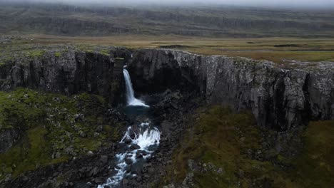 Snaedalsfoss-Wasserfall-Von-Der-Klippe-In-Bewölkter,-Düsterer-Isländischer-Landschaft,-Luftaufnahme