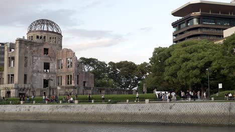 Cúpula-De-La-Bomba-Atómica-En-Hiroshima-Vista-Desde-El-Otro-Lado-Del-Río-Motoyasu,-Gente-Caminando-Por-El-Parque