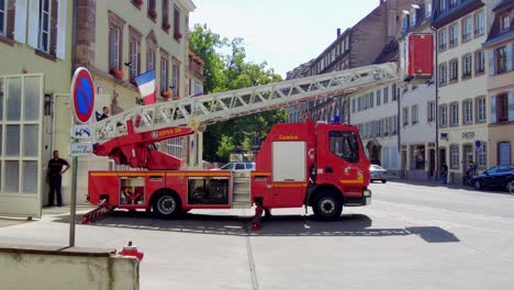 Französisches-Feuerwehrauto,-Das-In-Einem-Dorf-Stationiert-Ist-Und-Die-Drehleiter-Bewegt,-Mit-Einer-Französischen-Flagge-Im-Hintergrund