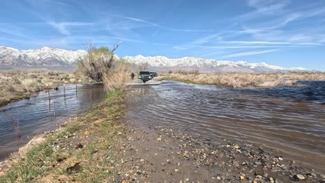 Snowy-Sierra-Nevada-mountain-range-with-a-bronco-driving-through-a-river-in-the-foreground-in-slow-motion