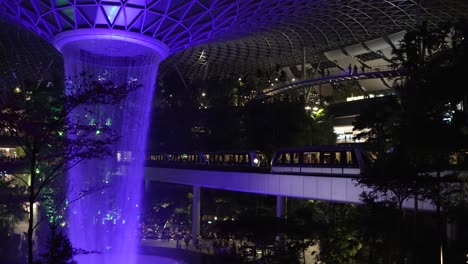 Cascada-Interior-Iluminada-De-Color-Púrpura-En-El-Aeropuerto-Jewel-Changi-De-Singapur-Por-La-Noche-Con-El-Skytrain-Pasando