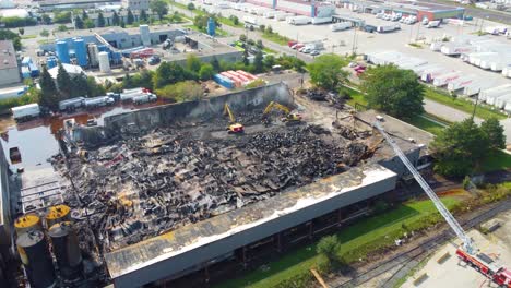 Disparo-Aéreo-De-Un-Dron-Descendiendo-Sobre-Los-Restos-De-Un-Depósito-De-Propano-Después-De-Un-Incendio-Industrial,-Mientras-Un-Equipo-De-Bomberos-Con-Excavadoras-Limpian-Los-Escombros-En-La-Devastadora-Escena,-Canadá