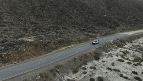 Aerial-drone-shot-of-over-a-speeding-white-car-on-a-highway-along-the-mountain-road-in-Hingol-Balochistan-during-evening-time