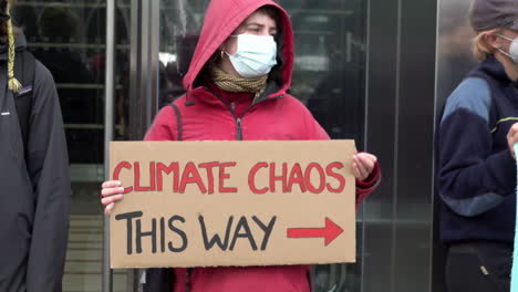 A-masked-protestor-in-a-red-hoody-holds-a-handmade-cardboard-placard-that-reads,-“Climate-chaos-this-way”
