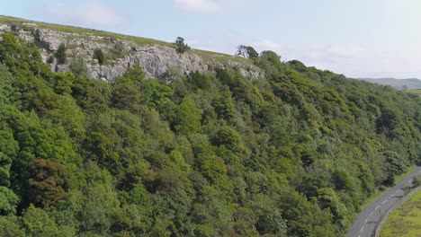 Drone-footage-from-a-rural-country-road-ascending-up-a-steep,-tree-lined-hilly-crag-revealing-the-Yorkshire-countryside,-farmland-and-fields,-dry-stone-walls-with-a-hilly-landscape-in-the-distance