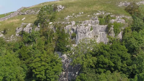 Drone-footage-ascending-up-and-over-a-steep,-tree-lined-limestone-crag-revealing-the-rural-Yorkshire-countryside-with-farmland,-fields,-dry-stone-walls-and-hilly-landscape-in-the-distance