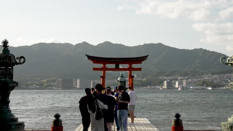 Visitors-Queuing-To-Take-Photo-Beside-Kadomarodo-Shrine-With-Floating-Grand-Torii-Gate-of-Itsukushima-Shrine-In-Background