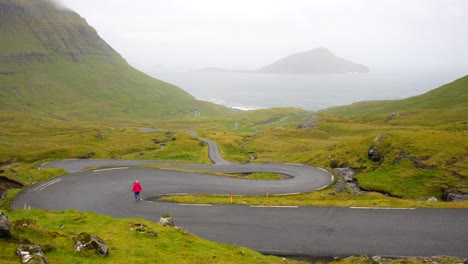 Female-hiker-walking-down-winding-Nordradalsskard-street,-Faroe-Islands