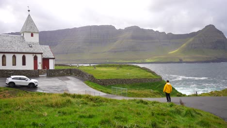 Male-tourist-walking-towards-car-near-white-church-of-Vidareidi-facing-Ocean,-Faroe-Islands