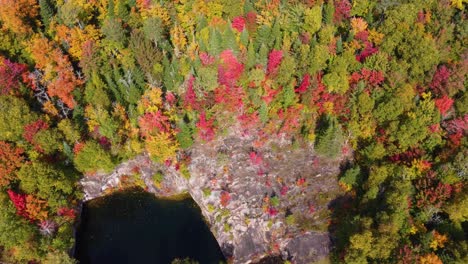 Aerial-drone-shot-descending-on-a-rock-face-of-a-lake-surrounded-by-a-forest-with-colorful-vibrant-autumn-colored-trees