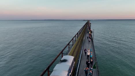 Die-Aufnahme-Der-Drohne-Bei-Sonnenuntergang-Am-Southend-Pier-Zeigt-Menschen,-Die-Am-Pier-Entlanggehen,-Während-Der-Pier-Zug-In-Die-Aufnahme-Eintritt-Und-Entlang-Der-Schienen-Fährt,-Sich-Dann-Zurückzieht-Und-Dem-Zug-Folgt