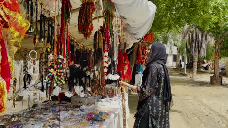 Mujer-Paquistaní-Con-Pañuelo-En-La-Cabeza-Caminando-Hacia-El-Puesto-De-Mercado-Vendiendo-Cuentas-Coloridas-En-Hingol,-Baluchistán
