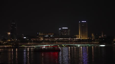 Night-time-cityscape-of-the-Marina-Bay-with-the-Jubilee-Bridge-in-Singapore-city,-and-transport-boats-passing-by