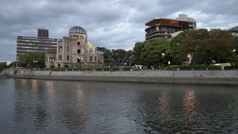 The-Atomic-Bomb-dome-building-with-people-walking-in-front-of-the-Motoyasu-River,-cloudy-evening