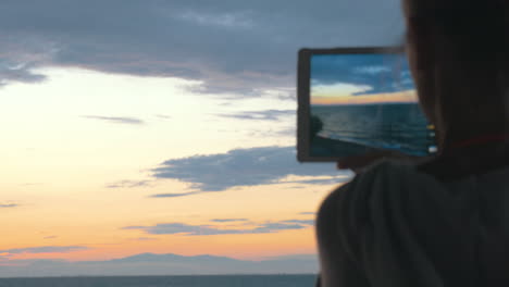Woman-takes-photos-of-the-beautiful-scenery-of-the-sea-and-evening-sky-with-her-tablet-at-sunset