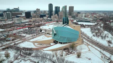 Establishing-Shot-Canadian-Museum-of-Human-Rights-Urban-Winnipeg-Manitoba-Canada-Downtown-Skyscraper-Buildings-in-City-Overcast-Landscape-Skyline-Snowing-Winter-Drone-4k-Shot-Multicolored-Train