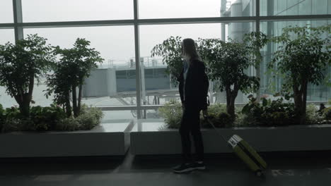 Girl-with-cell-and-suitcase-walking-by-the-window-of-airport-terminal
