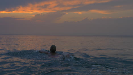 Young-woman-swims-away-in-the-sea-at-sunset-magical-view-of-evening-cloudy-sky-and-calm-sea