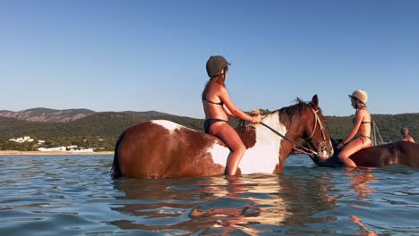 Young-girl-with-red-hair-and-cap-have-fun-bathing-while-riding-bareback-spotted-horse-in-sea-water-during-summer-season
