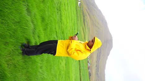 Man-wearing-yellow-raincoat-using-phone-in-Faroese-Vidareidi's-grass-field