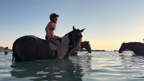 Cute-young-girl-with-equestrian-cap-and-group-of-people-riding-bareback-white-and-brown-horse-in-sea-water-during-in-season-at-sunset