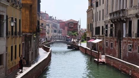 Una-Mujer-Camina-Por-El-Estrecho-Callejón-Vacío-Junto-Al-Canal-Con-Un-Puente-En-Venecia,-Italia