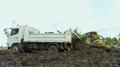 Dumping-some-more-soil-using-a-dump-tuck-and-a-grader-machine-in-an-arable-farm-land-to-get-it-ready-for-planting,-in-Chachoengsao-province-in-Thailand