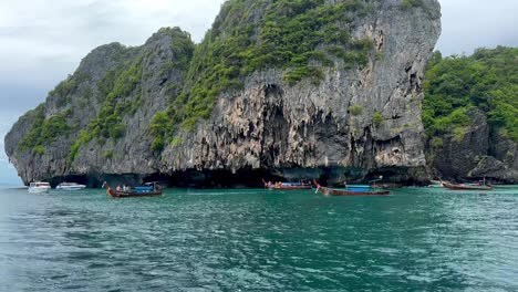 A-captivating-scene-near-Phi-Phi-Island,-Thailand:-a-majestic-rock-adorned-with-lush-greenery,-framed-by-turquoise-waters-where-four-long-tail-boats-gracefully-navigate