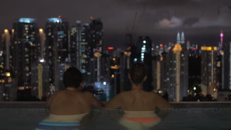 En-La-Piscina-En-La-Azotea-De-Un-Hotel-En-Kuala-Lumpur,-Malasia,-Un-Hombre-Con-Una-Mujer-Mirando-A-La-Ciudad-De-Noche.