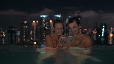 Vista-Nocturna-De-Una-Joven-Y-Hermosa-Pareja-En-La-Piscina-Mirando-En-Una-Tableta-En-El-Techo-Del-Rascacielos-Y-El-Paisaje-De-La-Ciudad-Kuala-Lumpur-Malasia