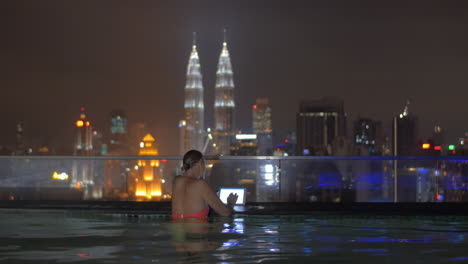 Blick-Auf-Eine-Frau-Im-Schwimmbad-Auf-Dem-Dach-Des-Wolkenkratzers-Mit-Tablet-Vor-Der-Nächtlichen-Stadtlandschaft-Von-Kuala-Lumpur,-Malaysia