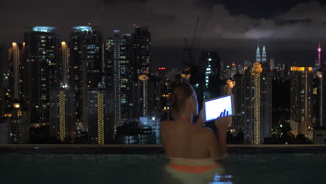 Blick-Auf-Eine-Frau,-Die-Im-Schwimmbad-Auf-Dem-Wolkenkratzerdach-Notizen-Mit-Einem-Tablet-Macht,-Und-Die-Nächtliche-Stadtlandschaft-Von-Kuala-Lumpur,-Malaysia