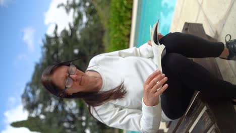 Vertical-View-Of-An-Adult-Woman-Wearing-Glasses-Is-Reading-Book-On-The-Poolside
