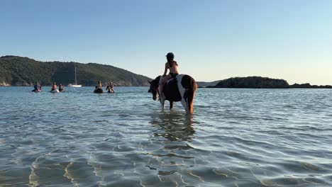 Young-girl-with-equestrian-helmet-have-fun-bathing-while-riding-bareback-white-and-brown-horse-in-ocean-water-during-summer-season-at-sunset