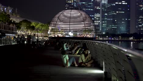 La-Gente-Se-Sentaba-Junto-A-La-Barandilla-Relajándose-En-El-Paseo-Marítimo-Junto-Al-Hotel-Marina-Bay-Sands-Con-Vistas-A-La-Apple-Store-Al-Fondo-Por-La-Noche-En-Singapur