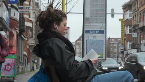 Young-woman-takes-time-to-read-and-listen-to-music-while-waiting-on-bus