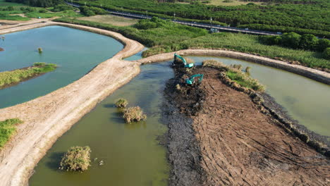 Excavator-on-muddy-dirt-island-restores-wetland-in-Guandu-Nature-park,-aerial-overview