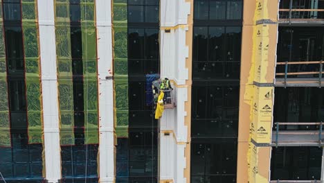Aerial-orbit-shot-showing-worker-in-lift-working-on-facade-of-skyscraper-tower-at-construction-site