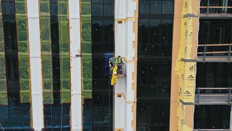 Toma-Aérea-En-órbita-Que-Muestra-A-Un-Trabajador-En-Un-Ascensor-Trabajando-En-La-Fachada-De-Una-Torre-De-Rascacielos-En-El-Sitio-De-Construcción.