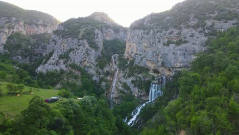 Aerial-backward-view-of-Ujevara-E-Sotires-wild-waterfalls-in-Nivica-Canyon,-with-cliffs-bathed-in-the-warm-hues-of-a-stunning-sunrise-during-the-golden-hour