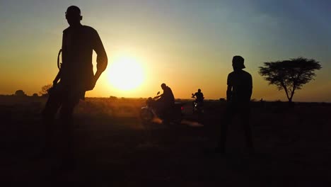 Silhouette-Ugandischer-Männer,-Die-Während-Des-Sonnenuntergangs-Spaß-Daran-Haben,-Mit-Fahrern-Auf-Motorrädern-Im-Hintergrund-Zu-Tanzen
