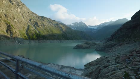 Pan-Left-View-Across-Calm-Lake-Alpe-Gera-Dam-With-Male-Hiker-With-Backpack-Walking-Past