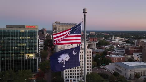 United-States-and-South-Carolina-flags-waving-proudly-over-Columbia,-SC-skyline-during-sunrise