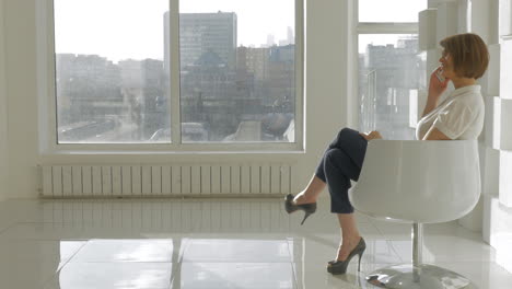 View-of-stylish-woman-sits-on-the-white-chair-and-haves-phone-call-against-window-with-cityscape-background