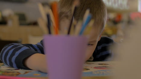 Kleiner-Junge-Löst-Kinderübung-Beim-Entwickeln-Eines-Buches-Für-Kinder-Im-Vorschulalter,-Unscharfes-Glas-Mit-Bleistiften-Im-Vordergrund