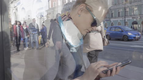 Blonde-Frau-Benutzt-Mobiltelefon-Vor-Glasreflexion-Von-Menschen,-Die-Sich-Auf-Der-Straße-Bewegen